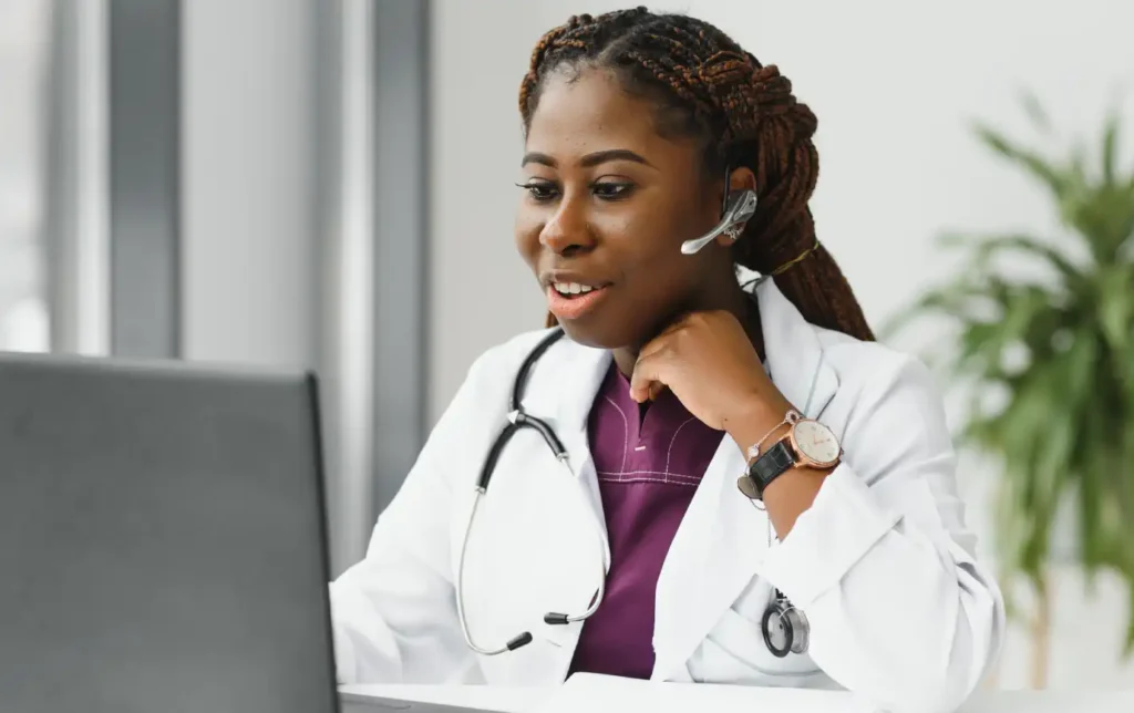 Médica sorridente usando headset, assistindo a um computador, em ambiente de trabalho moderno. Ideal para consultas médicas online e telemedicina.