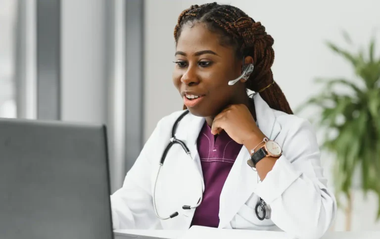 Médica sorridente usando headset, assistindo a um computador, em ambiente de trabalho moderno. Ideal para consultas médicas online e telemedicina.