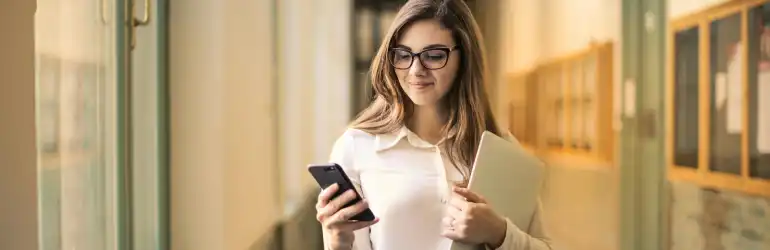 Inscrição no Revalida medicina: mulher jovem com óculos, checando o celular enquanto segura um notebook, em ambiente iluminado com paredes de vidro.