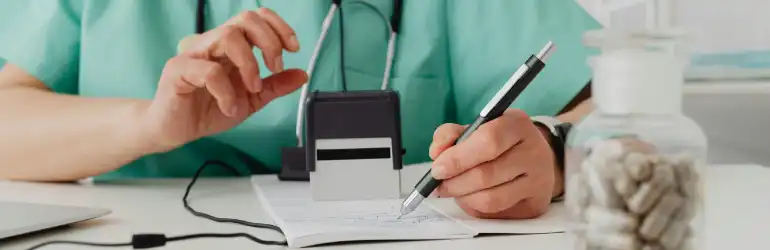 Médico de jaleco escrevendo em uma folha enquanto utiliza um estetoscópio e tem um vidro de cápsulas ao lado.