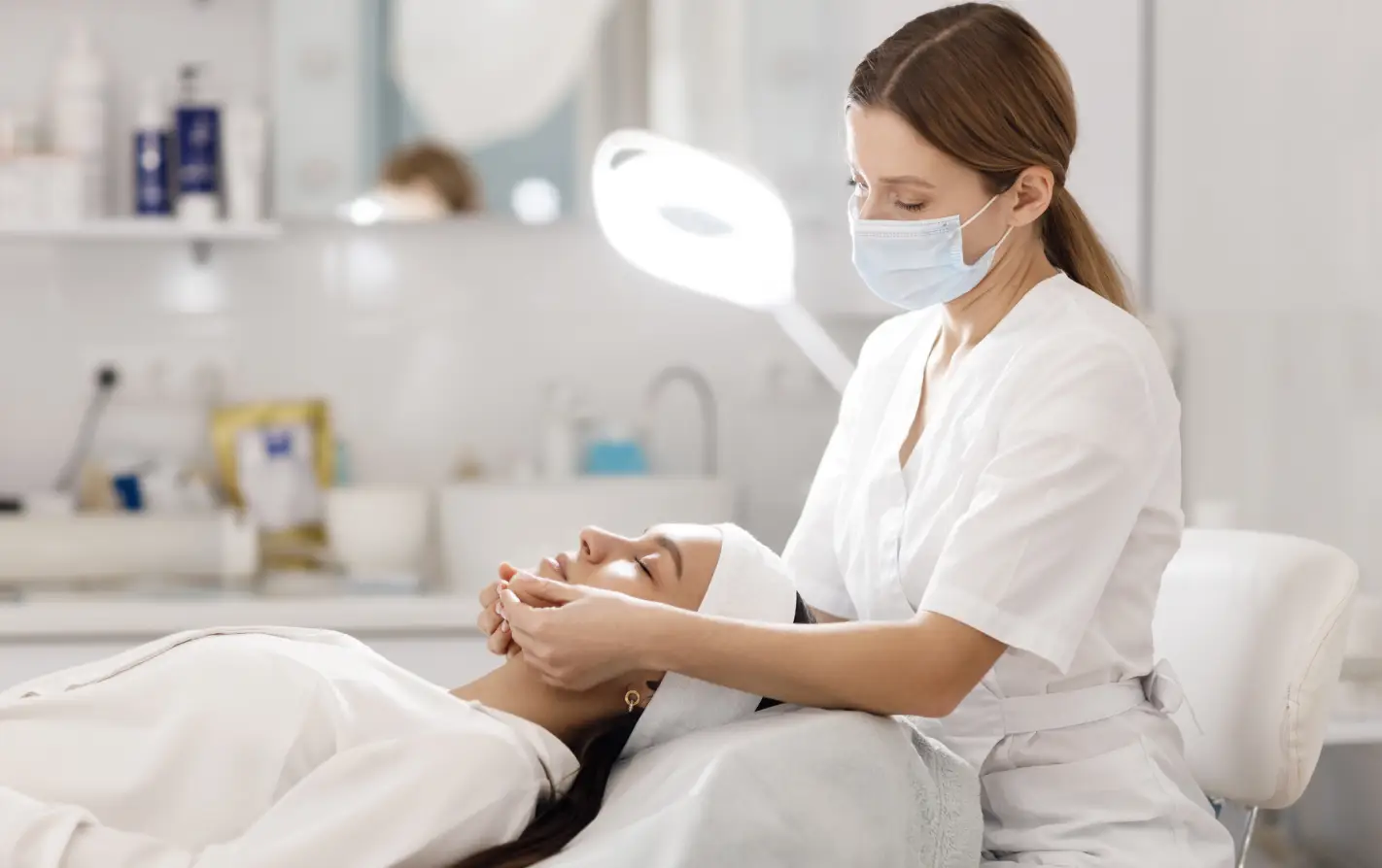 Uma esteticista de jaleco branco e máscara de proteção realiza um procedimento facial em uma cliente deitada na maca. A cena, iluminada por uma luminária de LED, ilustra o processo prático da anamnese estética em uma clínica de estética, mostrando a interação entre profissional e cliente antes de um tratamento.