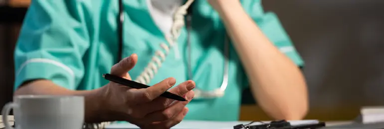 
Um profissional de enfermagem, vestindo um uniforme verde, está sentado em uma mesa e ao telefone. Ele segura uma caneta, com a outra mão apoiando o rosto. À frente, na mesa, há uma xícara branca e papéis, simbolizando a anamnese na enfermagem e o fluxo de informações que precisam ser organizados.