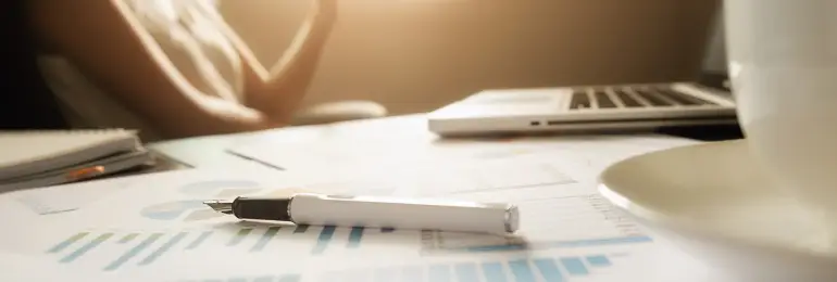 Imagem de uma mesa de trabalho com papel, caneta, laptop e xícara, ideal para estudos ou reuniões de negócios. Ambiente profissional e organizado.