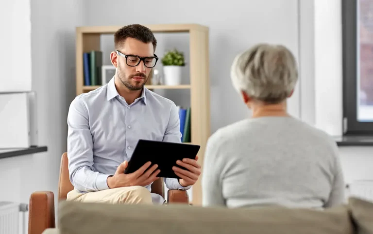 Um psicólogo de camisa social e óculos está sentado em um consultório, olhando para um tablet enquanto interage com uma paciente (vista de costas). A cena ilustra o profissional realizando a coleta de dados e informações essenciais, caracterizando a anamnese psicológica para a melhoria do atendimento clínico.