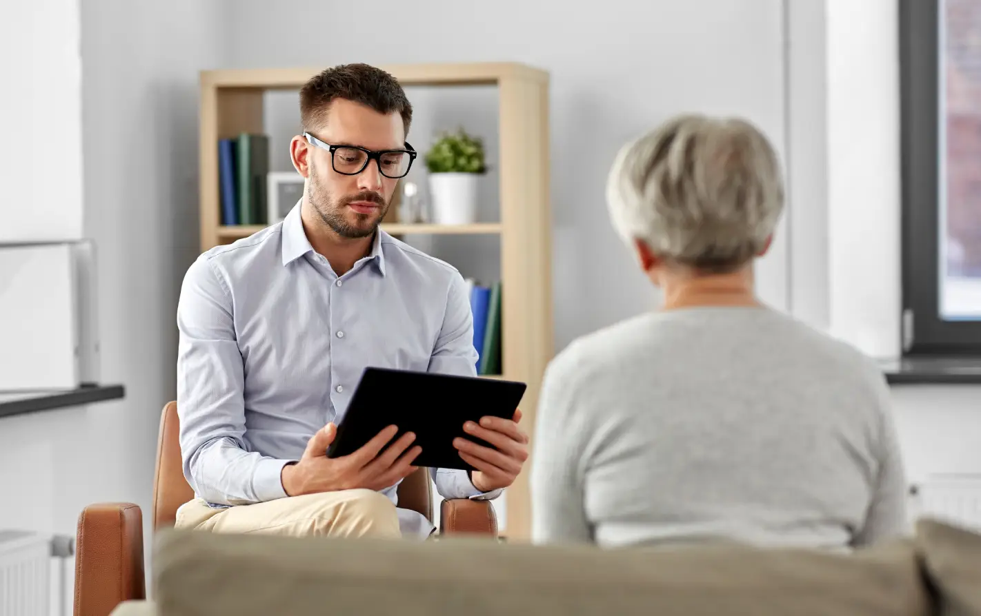 Um psicólogo de camisa social e óculos está sentado em um consultório, olhando para um tablet enquanto interage com uma paciente (vista de costas). A cena ilustra o profissional realizando a coleta de dados e informações essenciais, caracterizando a anamnese psicológica para a melhoria do atendimento clínico.