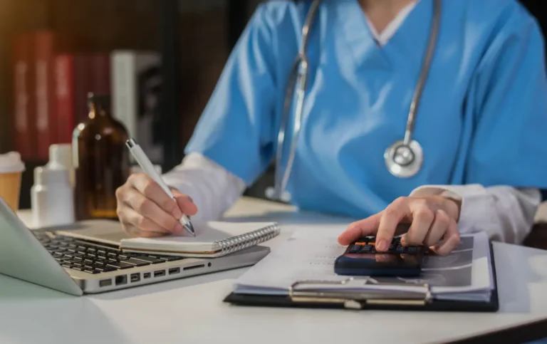 Um profissional de saúde, vestindo uniforme azul e estetoscópio, está em uma mesa, fazendo anotações em um caderno, usando um laptop e uma calculadora. A imagem ilustra as tarefas administrativas e financeiras em um consultório ou clínica, representando a importância do correto preenchimento da DMED (Declaração de Serviços Médicos e de Saúde) para a Receita Federal.
