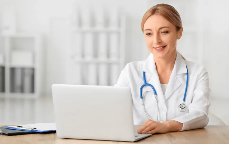 Uma médica sorridente, de jaleco branco e estetoscópio azul no pescoço, está sentada à sua mesa, trabalhando em um notebook. A imagem ilustra um profissional de saúde no contexto administrativo, destacando a necessidade de padronização e o uso do código TUSS para procedimentos médicos.