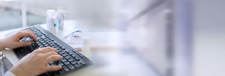 Mãos digitando em um teclado de computador em um ambiente de escritório médico. A imagem, com elementos de fundo desfocados, como frascos de medicamento, ilustra o trabalho manual e repetitivo, destacando os desafios e a complexidade do gerenciamento da guia SADT para faturamento e procedimentos.