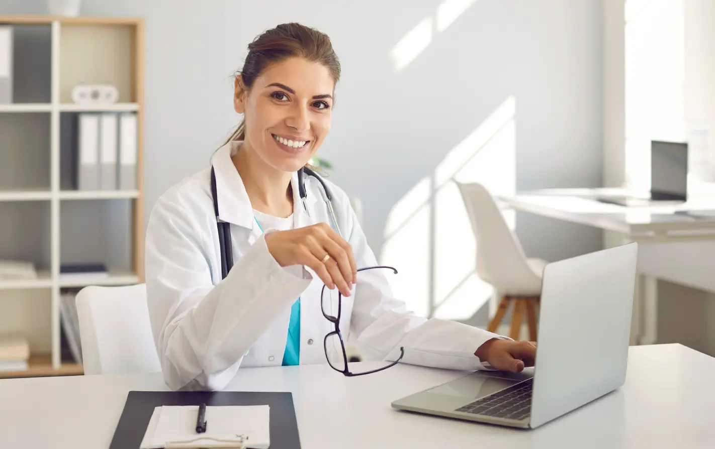 Uma médica sorridente, de jaleco branco e estetoscópio, está sentada em sua mesa de consultório com um notebook e segura seus óculos. A cena ilustra o profissional de saúde realizando tarefas administrativas e de faturamento, destacando a emissão e a função da guia SADT para solicitação de exames e procedimentos médicos.