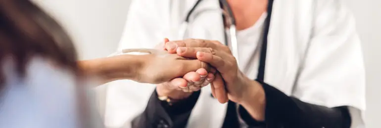 Mãos de um profissional de saúde, vestindo jaleco, segurando as mãos de um paciente, simbolizando o cuidado, a empatia e a importância do atendimento humanizado na saúde.