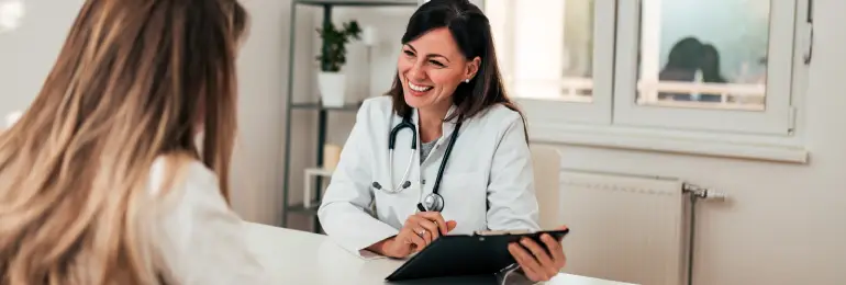 Médica sorridente em consulta, segurando uma prancheta enquanto interage com uma paciente, simbolizando a comunicação clara e os próximos passos no cuidado, destacando a importância do atendimento humanizado na saúde para o aprimoramento contínuo.