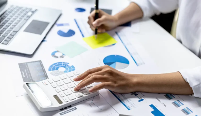 Gestor médico analisando gráficos e relatórios financeiros em uma mesa de escritório, com as mãos usando um teclado e apontando para documentos com gráficos, números e indicadores.
