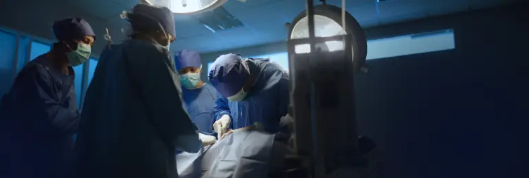 Equipe de cirurgiões em centro cirúrgico usando máscaras e roupas esterilizadas, realizando uma cirurgia com iluminação forte e instrumentos ao redor do paciente sob campo cirúrgico.