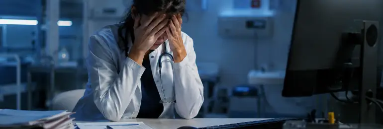 Médica com as mãos no rosto, expressão de cansaço e preocupação, usando jaleco branco e estetoscópio sobre uma mesa com prontuários e documentos.